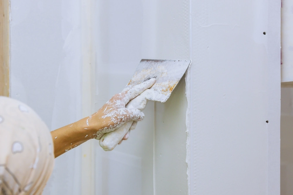 Hand mit Spachtel glättet weißen Putz auf Gipskartonwand im Rahmen der Spachtelarbeiten