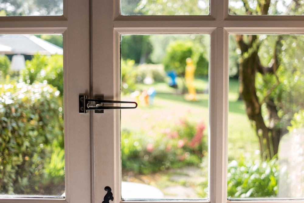 Weiße Holzfenster mit montiertem Sicherheitsriegel und unscharfem Gartenblick
